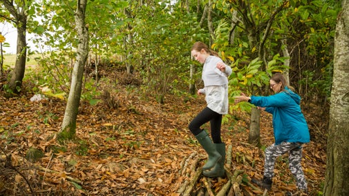 Woman and girl clambering uphill through woodland over logs and leaves